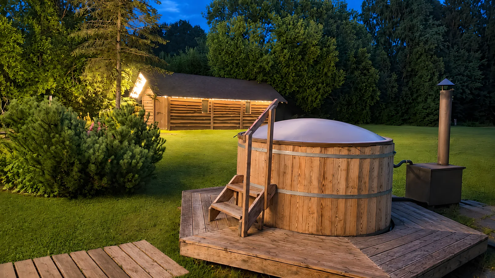 The hot tub on a wooden deck in the evening, its stove beside it and a lit outbuilding
