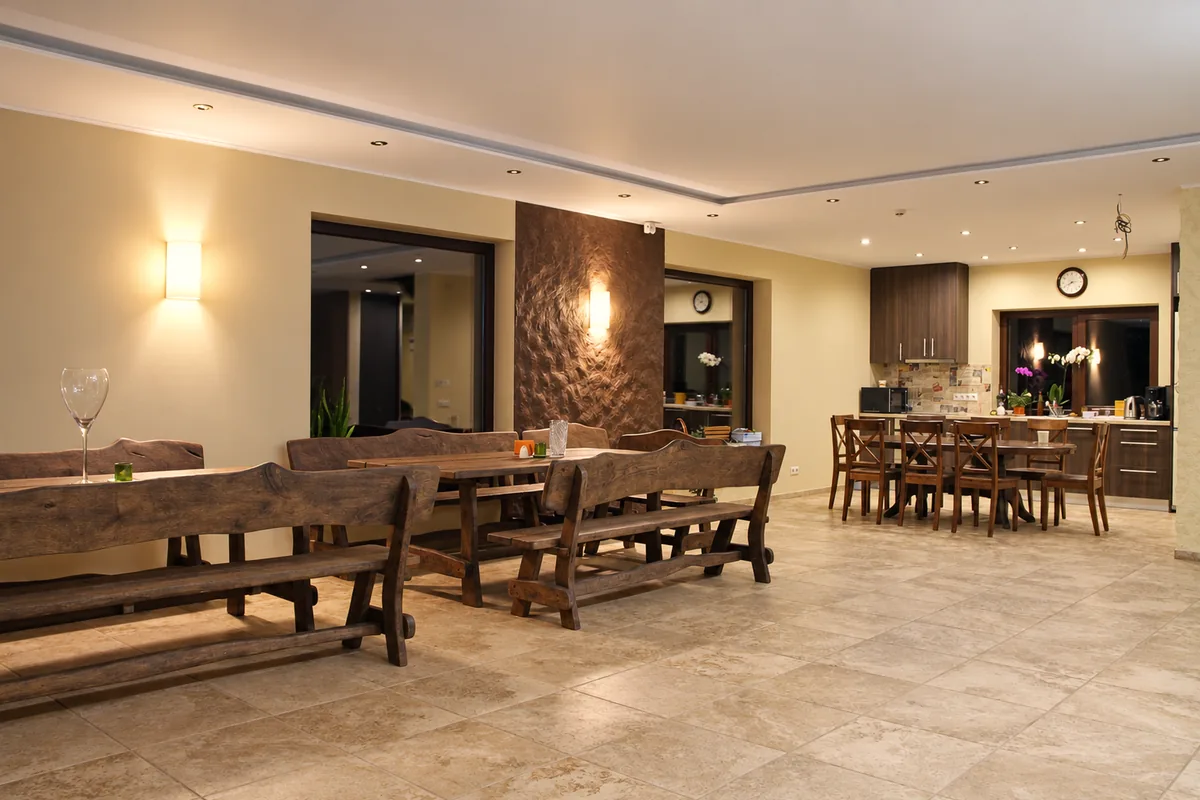 The hall in the evening light, long wooden tables and benches, the kitchen and a dining table beyond